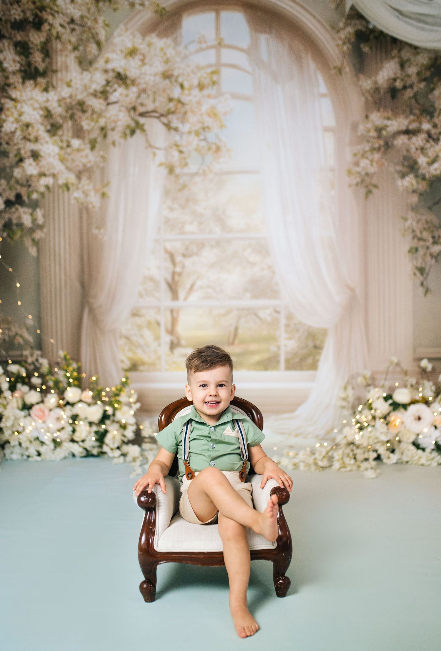 A smiling toddler boy in a green outfit sits in a wooden chair surrounded by white flowers and sheer curtains.