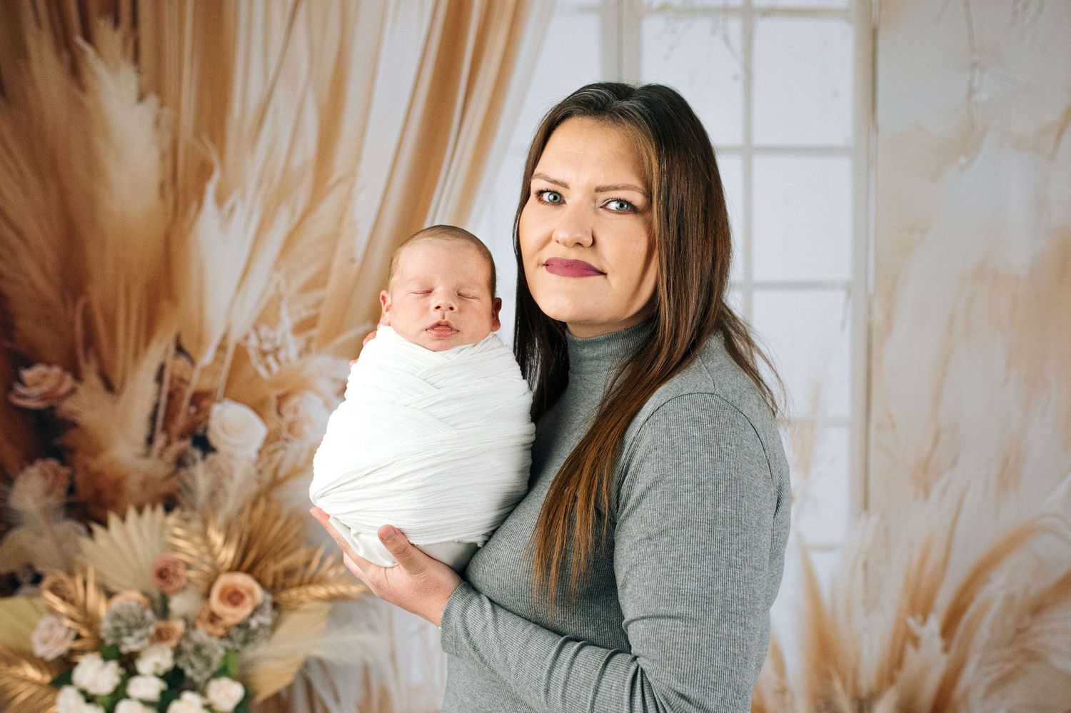 A woman in a gray turtleneck holds a sleeping newborn swaddled in white against a boho floral backdrop.
