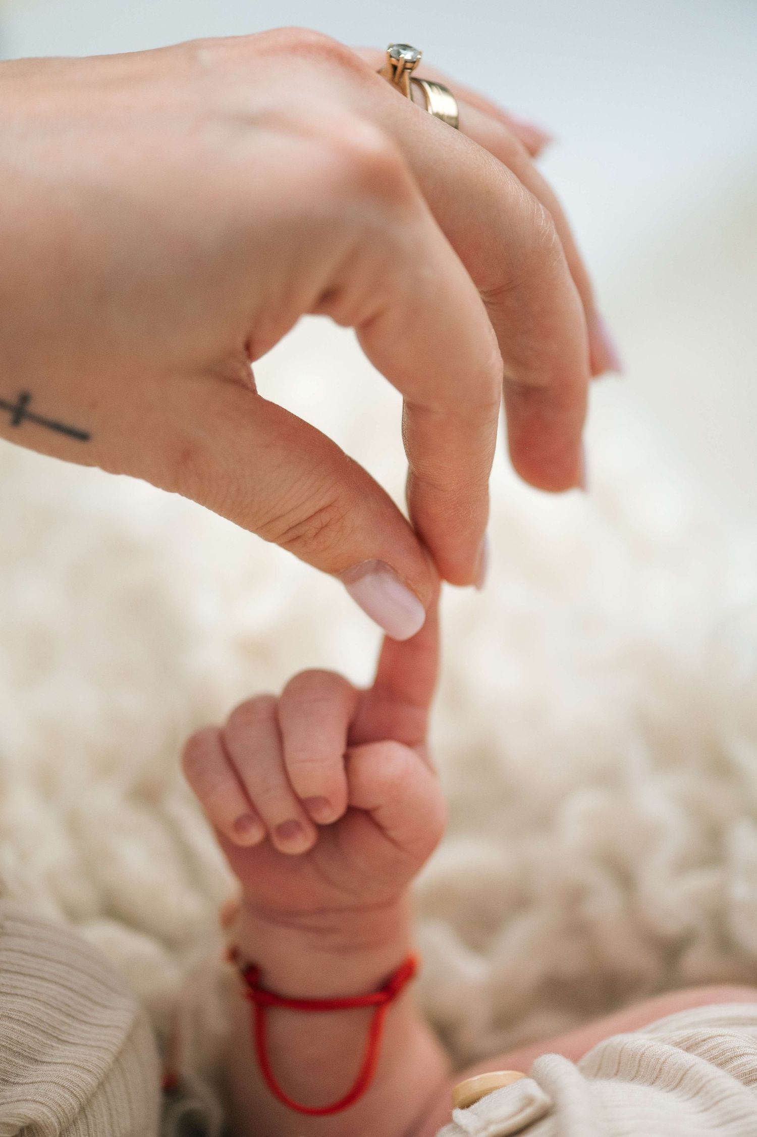A mother's hand with a gold ring and cross tattoo gently touching her newborn baby's tiny hand with a red bracelet.