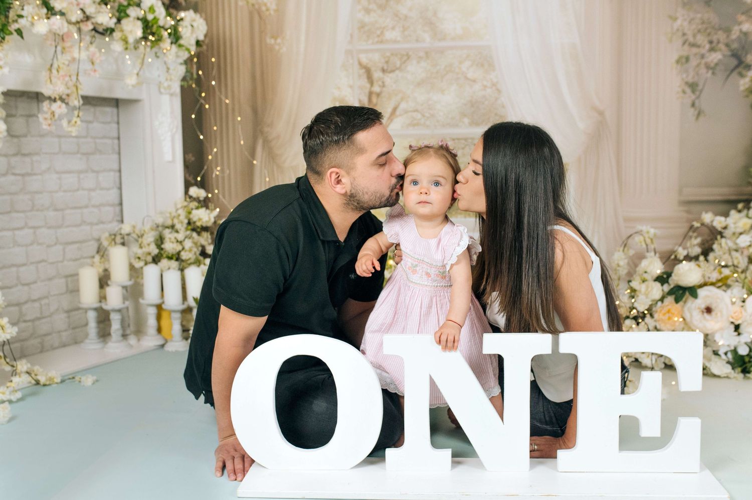 A family celebrates their baby girl's first birthday, with parents kissing her cheeks beside large ONE letters.