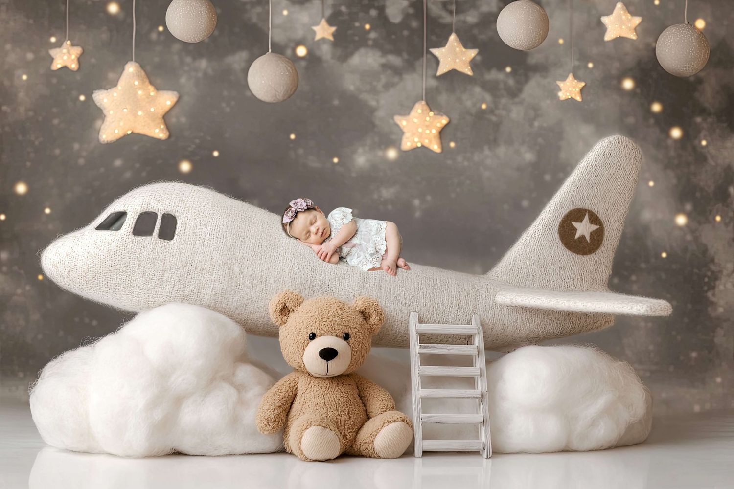 Newborn baby sleeping on a plush toy airplane with a teddy bear, stars, and clouds in a dreamy nursery photo.