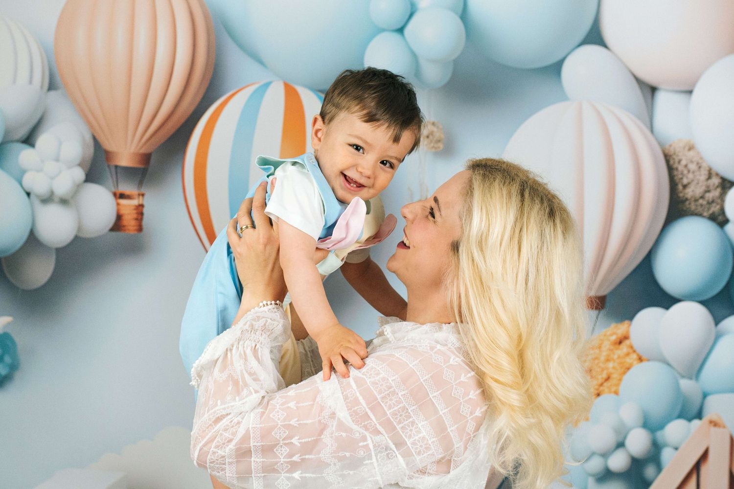 A blonde woman joyfully lifts a smiling baby boy surrounded by blue balloons and hot air balloon decorations.