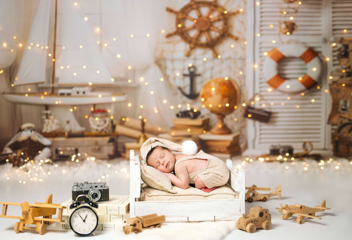 Newborn baby sleeping in white wooden crate surrounded by nautical decor, toy planes, clock, and fairy lights.
