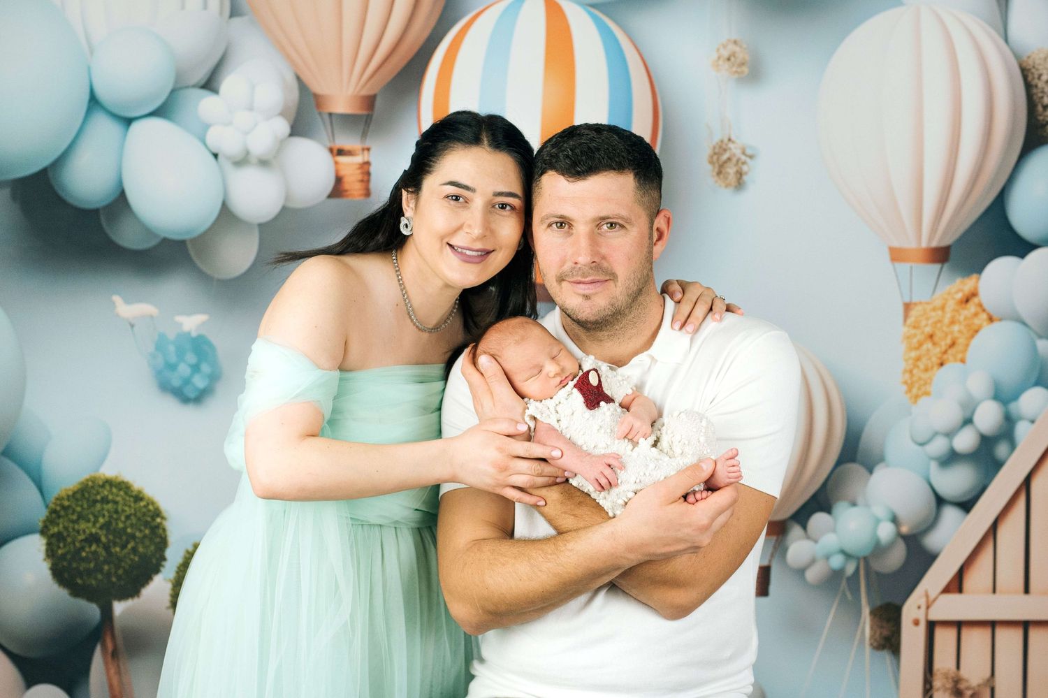 Happy family with newborn baby posing in front of blue balloon and hot air balloon decorated backdrop.