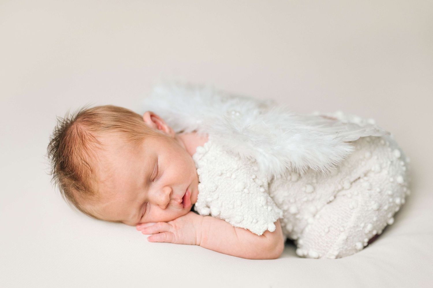 Sleeping newborn baby wrapped in white fluffy blanket with angel wings on white background.
