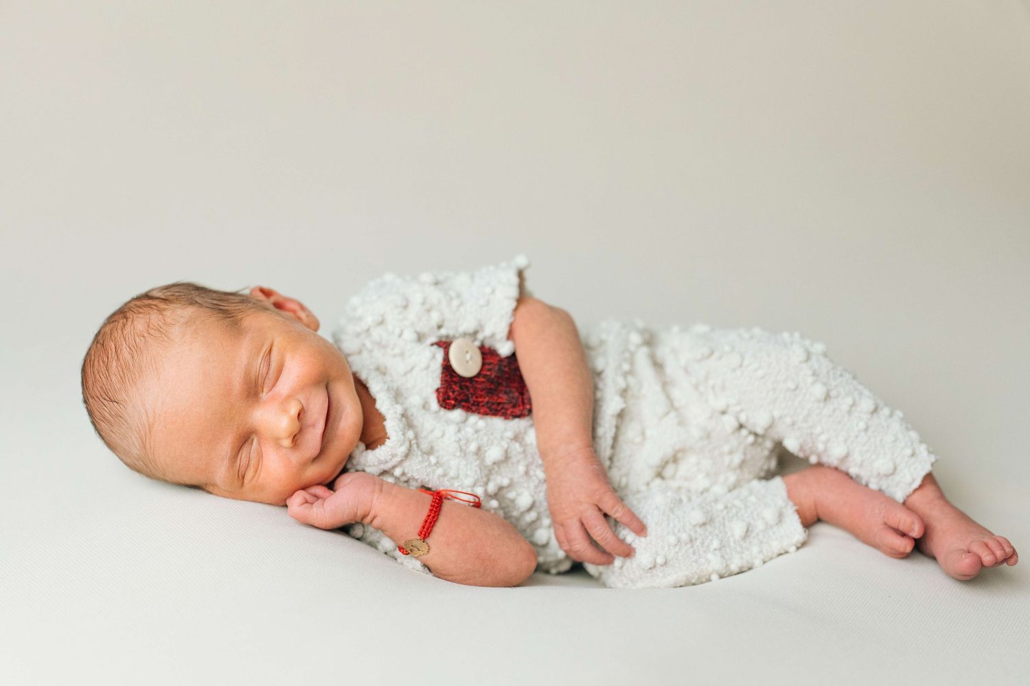 Sleeping newborn baby in white fuzzy outfit holding a small red toy on a light gray background.