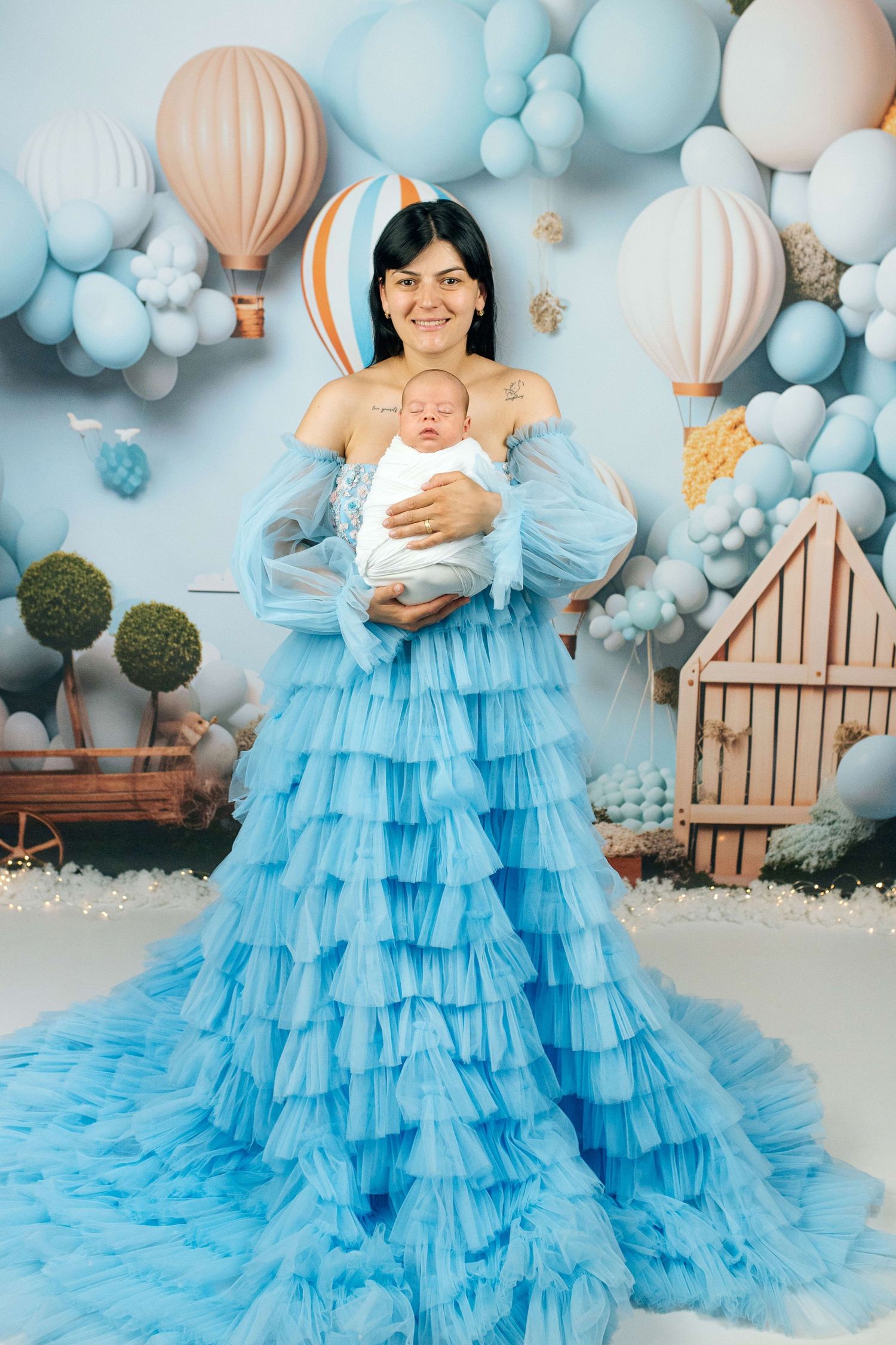 A woman in a blue ruffled gown holds a newborn baby, surrounded by blue balloons and hot air balloon decor.