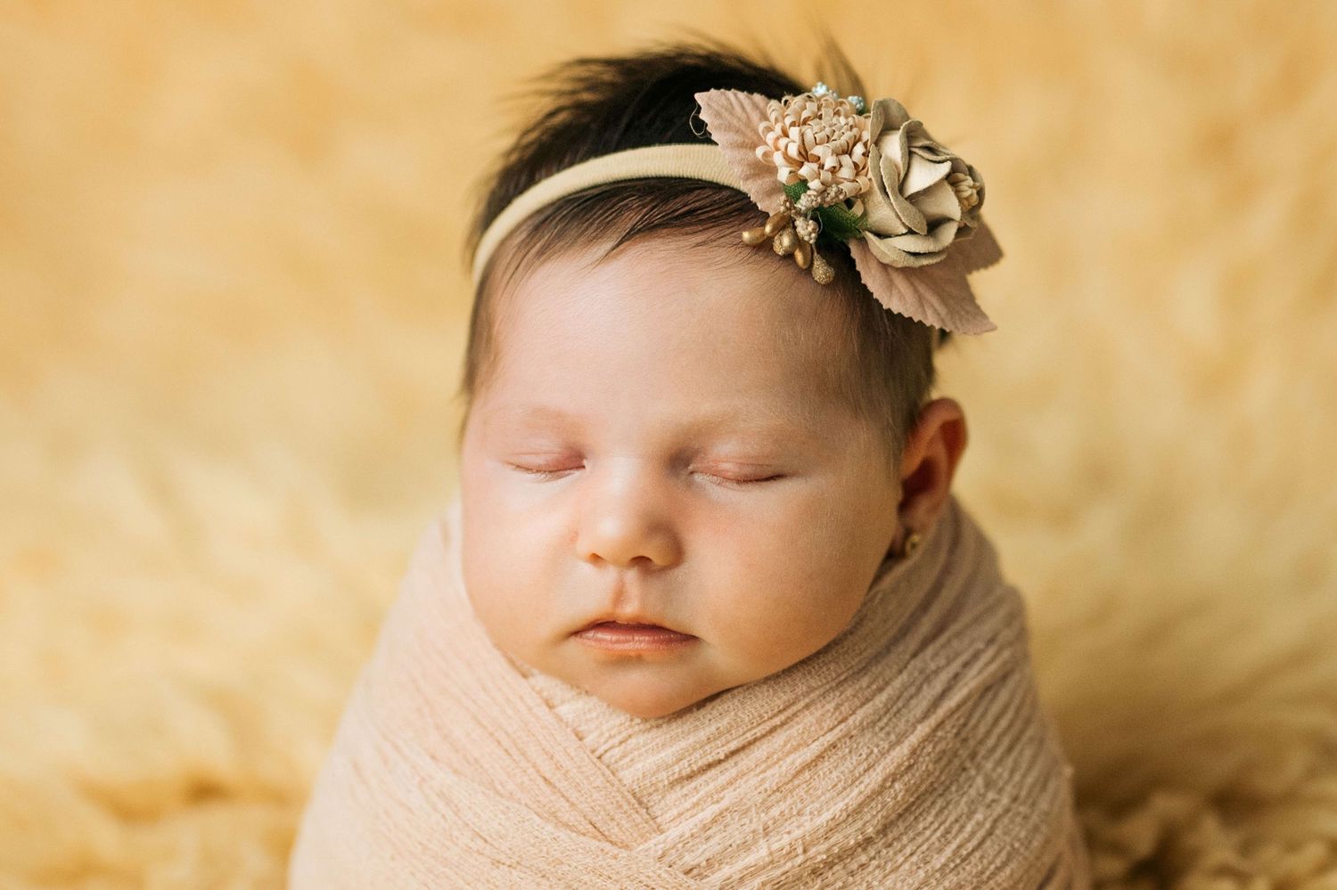 Sleeping newborn baby swaddled in beige wrap with floral headband on soft yellow background.
