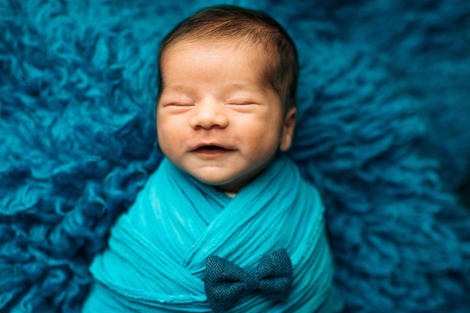 Smiling newborn baby wrapped in teal swaddle blanket with bow tie on fluffy blue background.