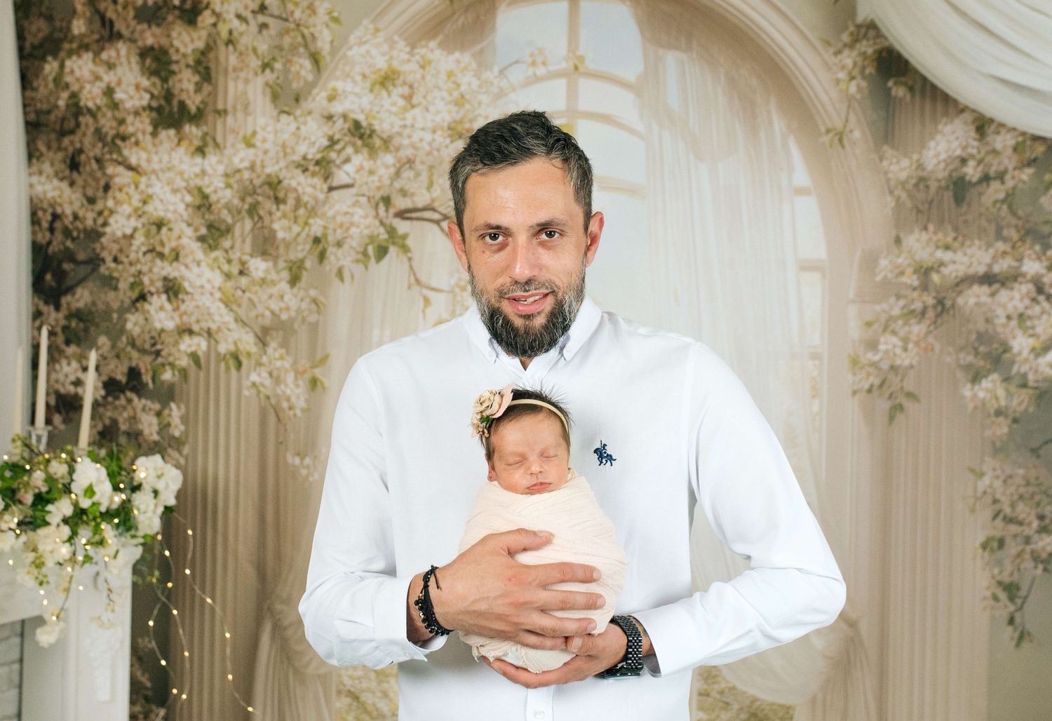 A man with a beard in a white shirt holds a newborn baby girl with a floral headband in an elegant floral setting.