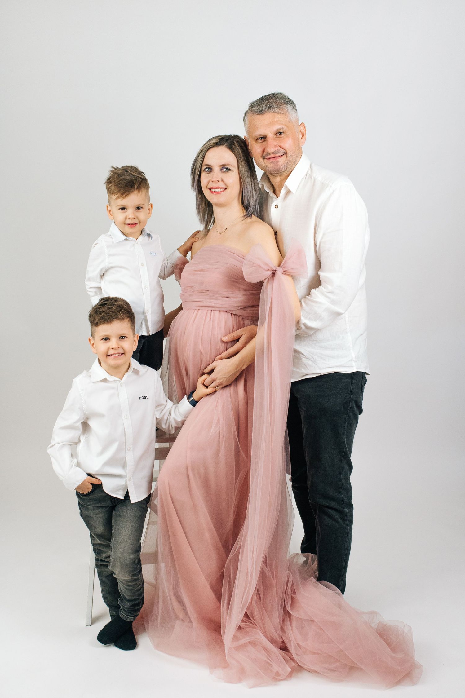 Pregnant woman in pink gown poses with husband and two young sons in white shirts for a family maternity photo.