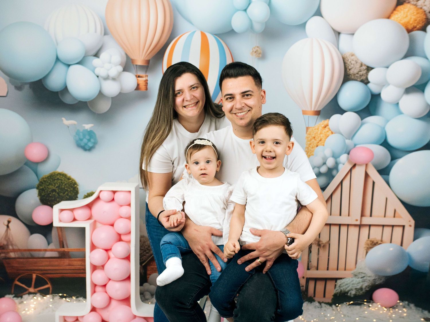 Family of four posing for a first birthday photo shoot with colorful balloons and hot air balloon backdrop.