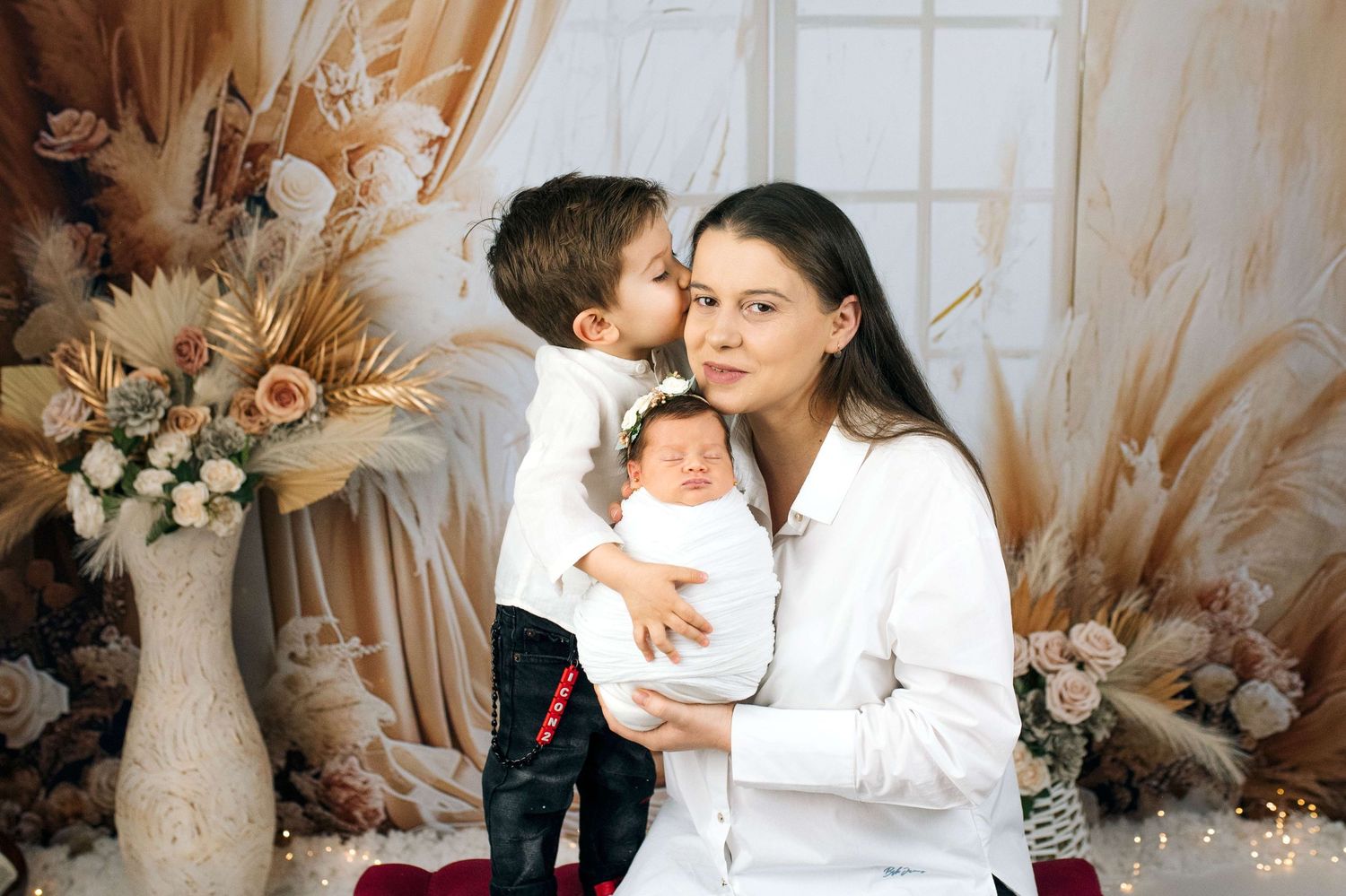 A mother in white holds her newborn while her toddler son gives her a sweet kiss on the cheek in a floral studio.