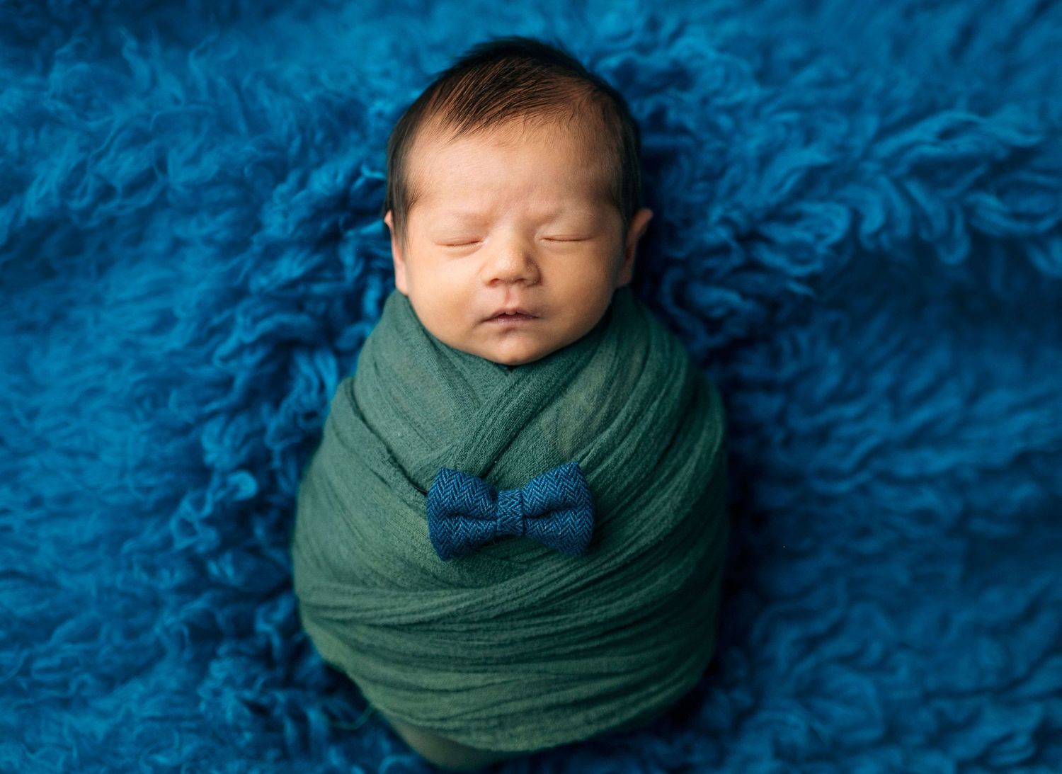Newborn baby swaddled in green wrap with blue bow tie, sleeping peacefully on a fluffy blue background.