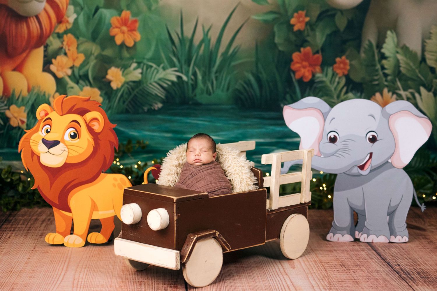 Newborn baby sleeping in a wooden toy car prop surrounded by cartoon lion and elephant in a jungle-themed photo shoot.
