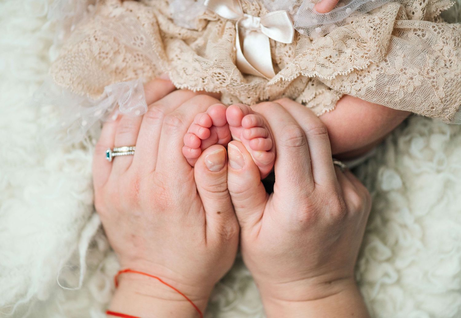 Adult hands gently cradling tiny newborn baby feet, with infant wearing delicate lace dress and bow.