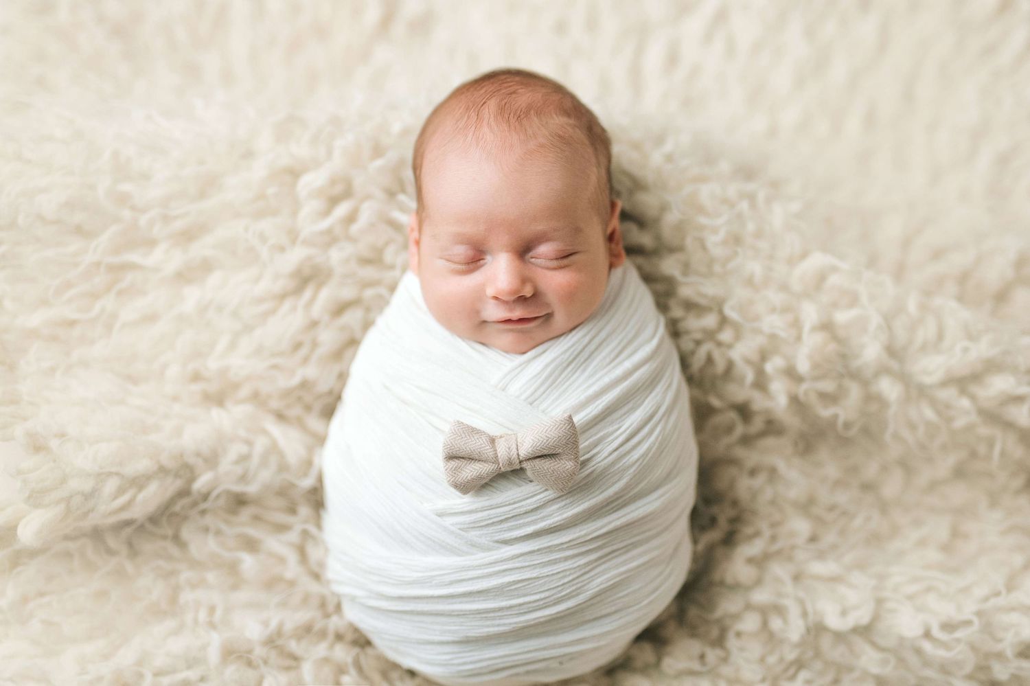 Smiling newborn baby swaddled in white wrap with bow tie on fluffy cream fur blanket.