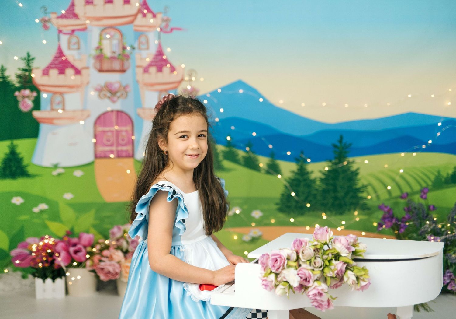 A young girl in a blue dress smiles in front of a fairy tale castle backdrop with flowers and twinkling lights.