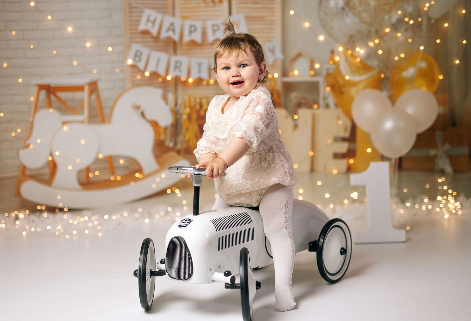 Smiling baby girl riding a white toy car at a first birthday party with golden decorations and fairy lights.