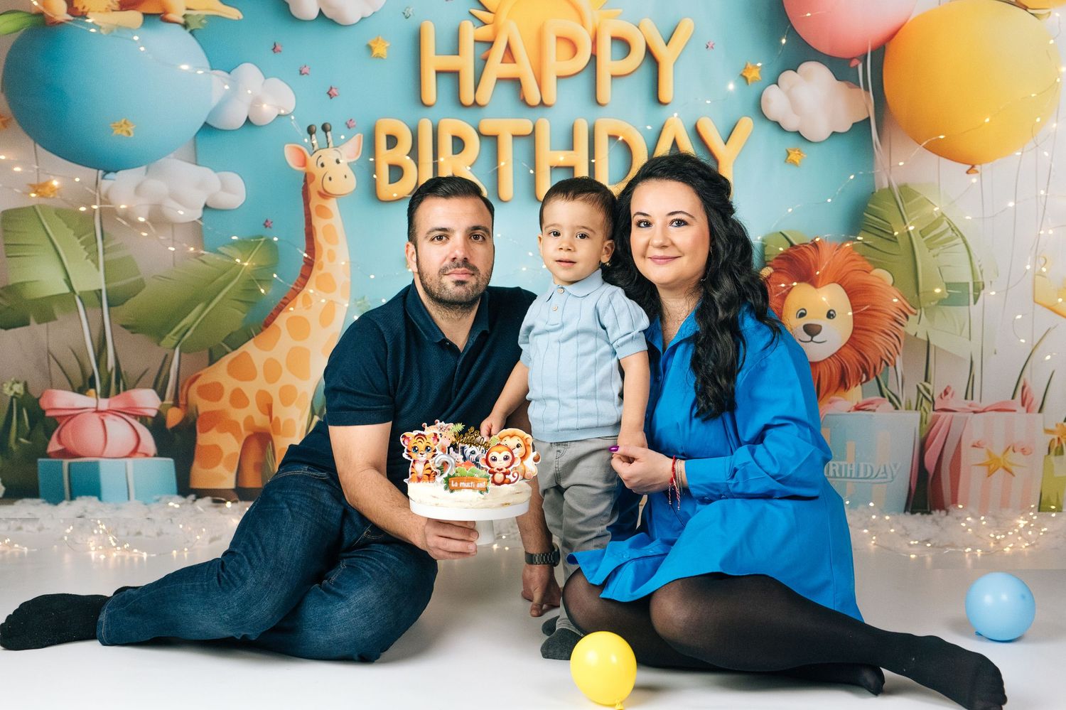 Family of three celebrating toddler's first birthday with cake in front of colorful Happy Birthday backdrop with animals.