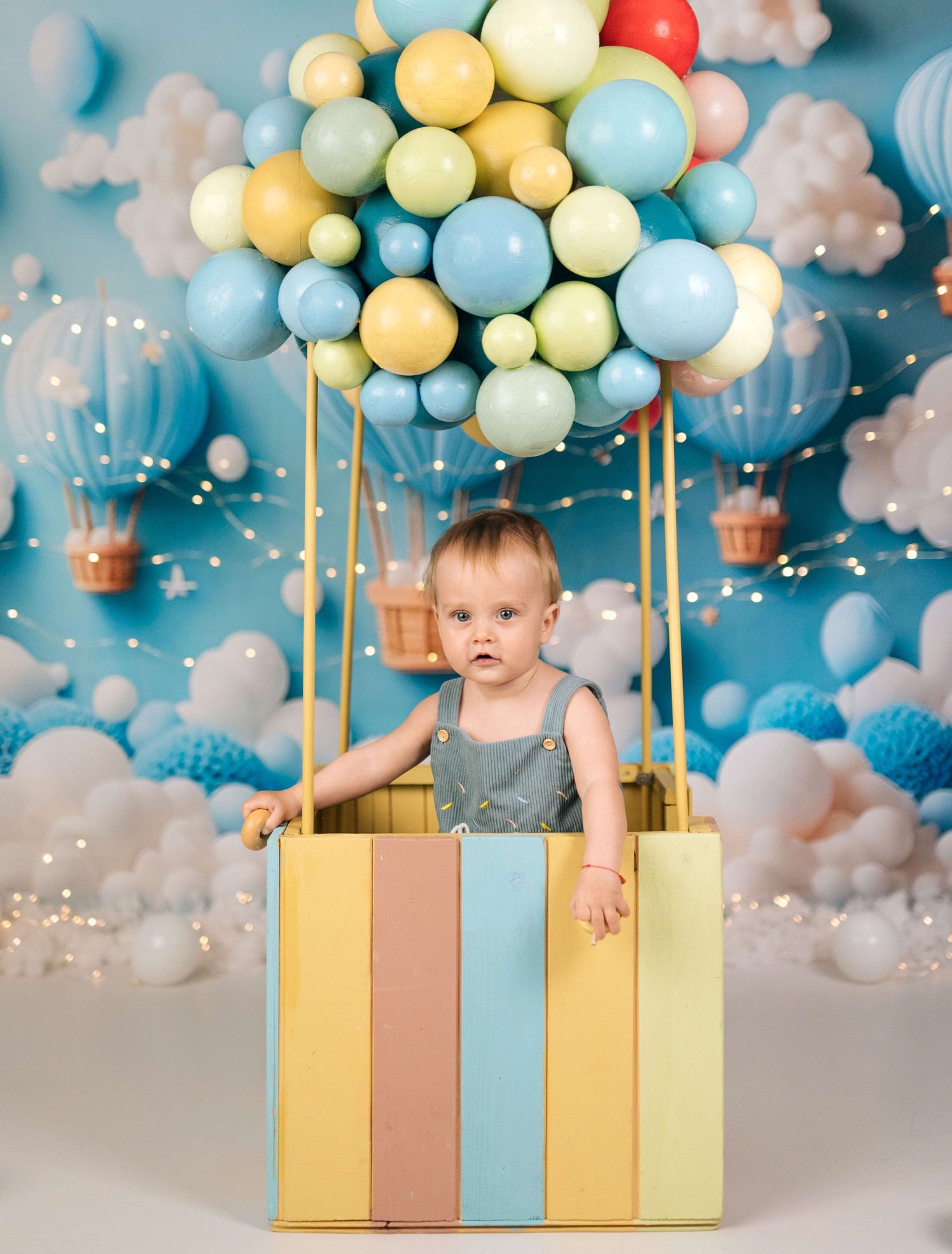 Baby sits in colorful striped box decorated as hot air balloon with pastel balloons on blue backdrop.
