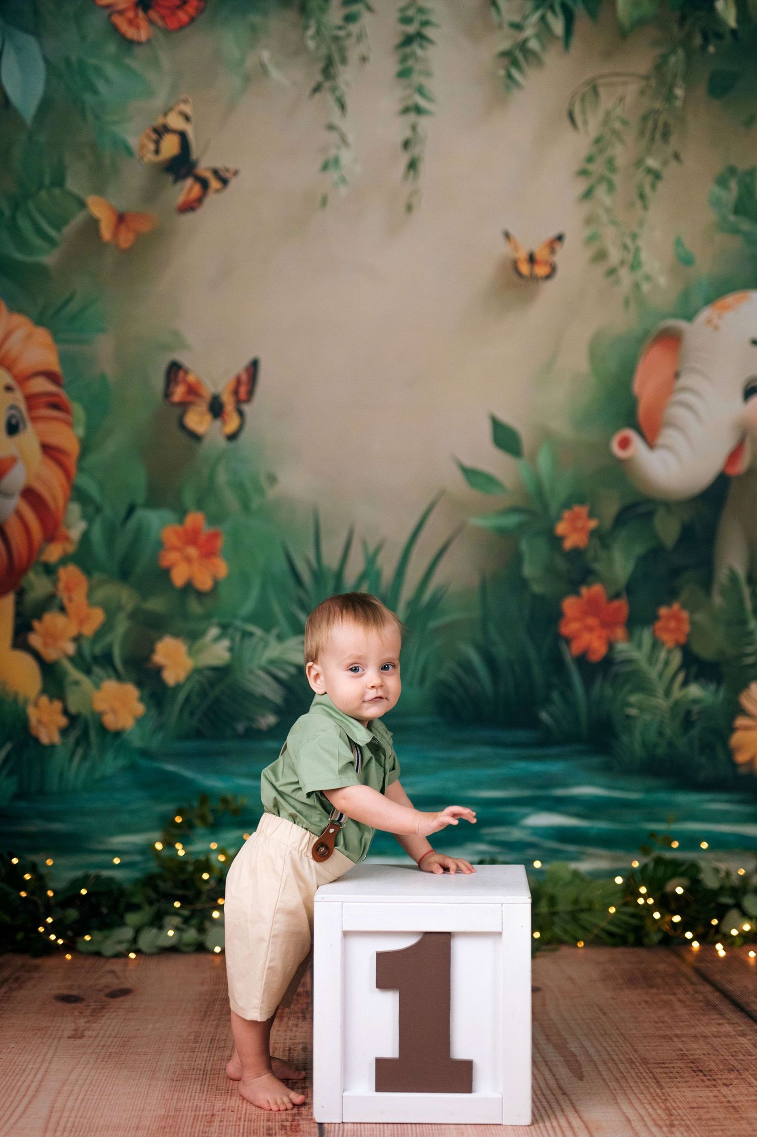Baby boy in green shirt stands by number 1 block, jungle safari backdrop with elephants and butterflies.