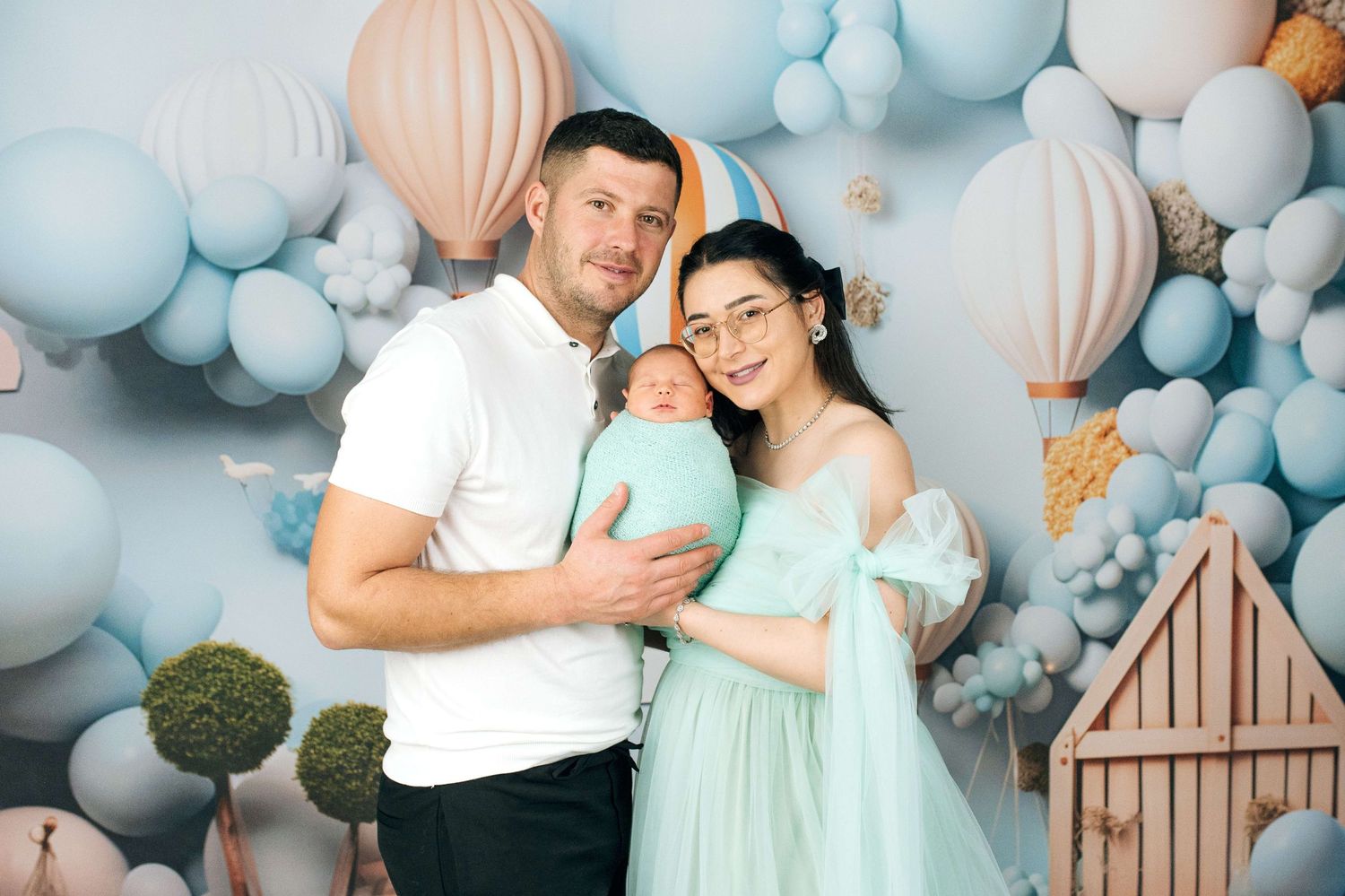 Young couple in mint outfits holding newborn baby in front of pastel blue balloon and hot air balloon backdrop.