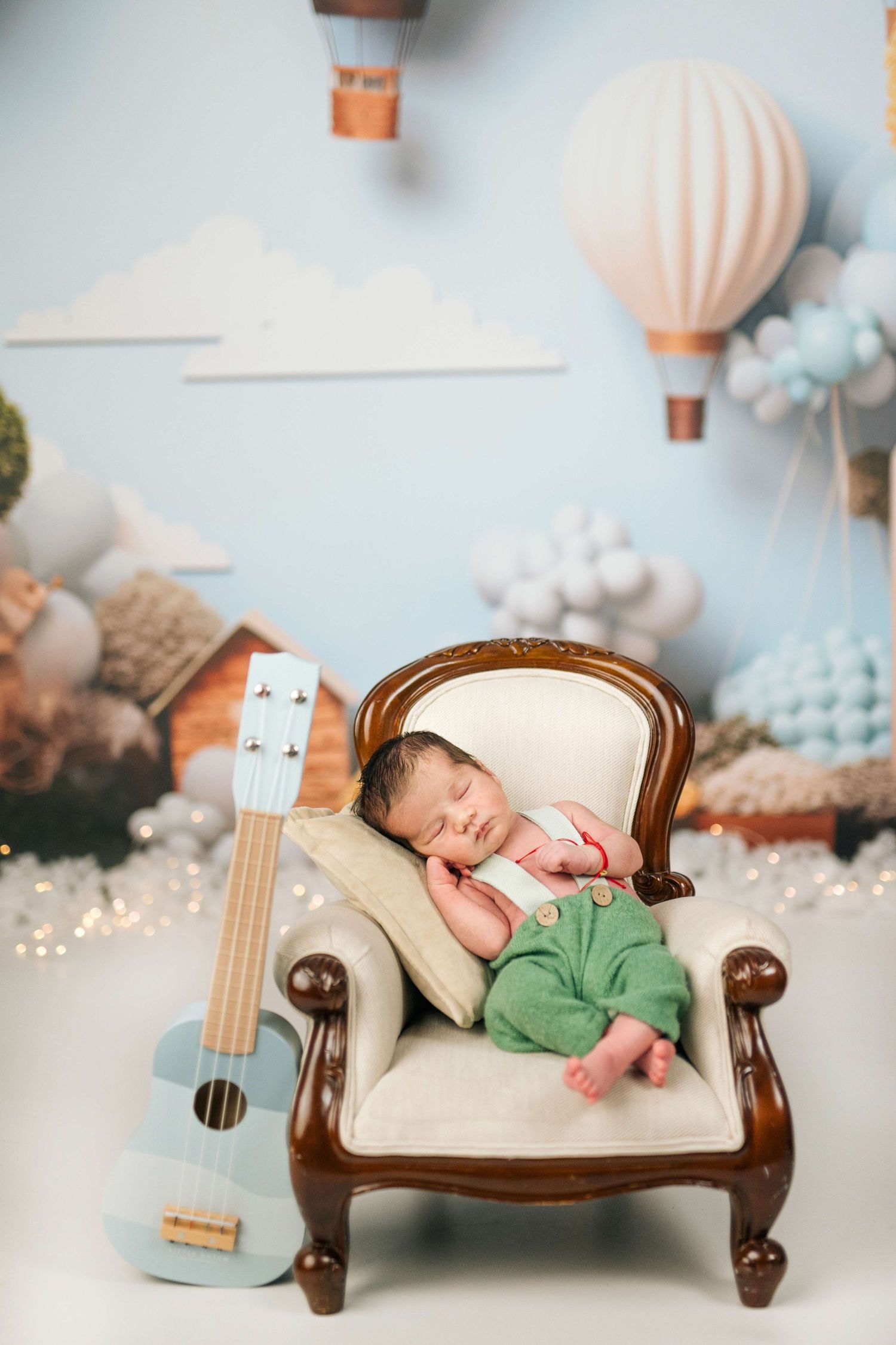 Newborn baby sleeping peacefully on a miniature armchair beside a blue ukulele, with hot air balloon backdrop.