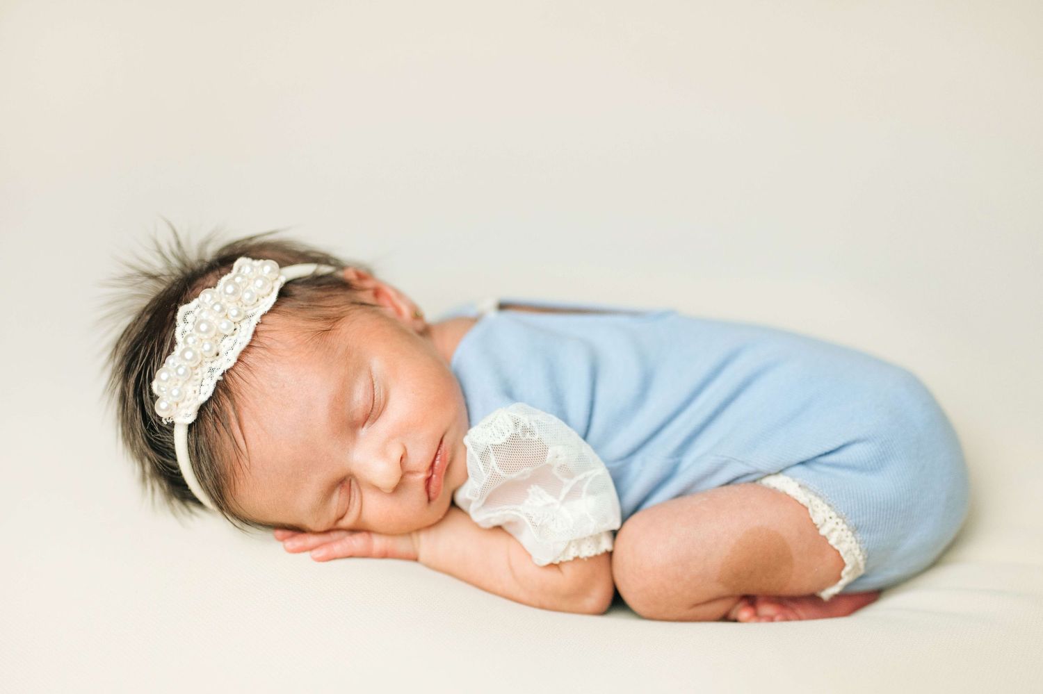 Newborn baby sleeping peacefully in blue outfit with white lace headband on soft white background.