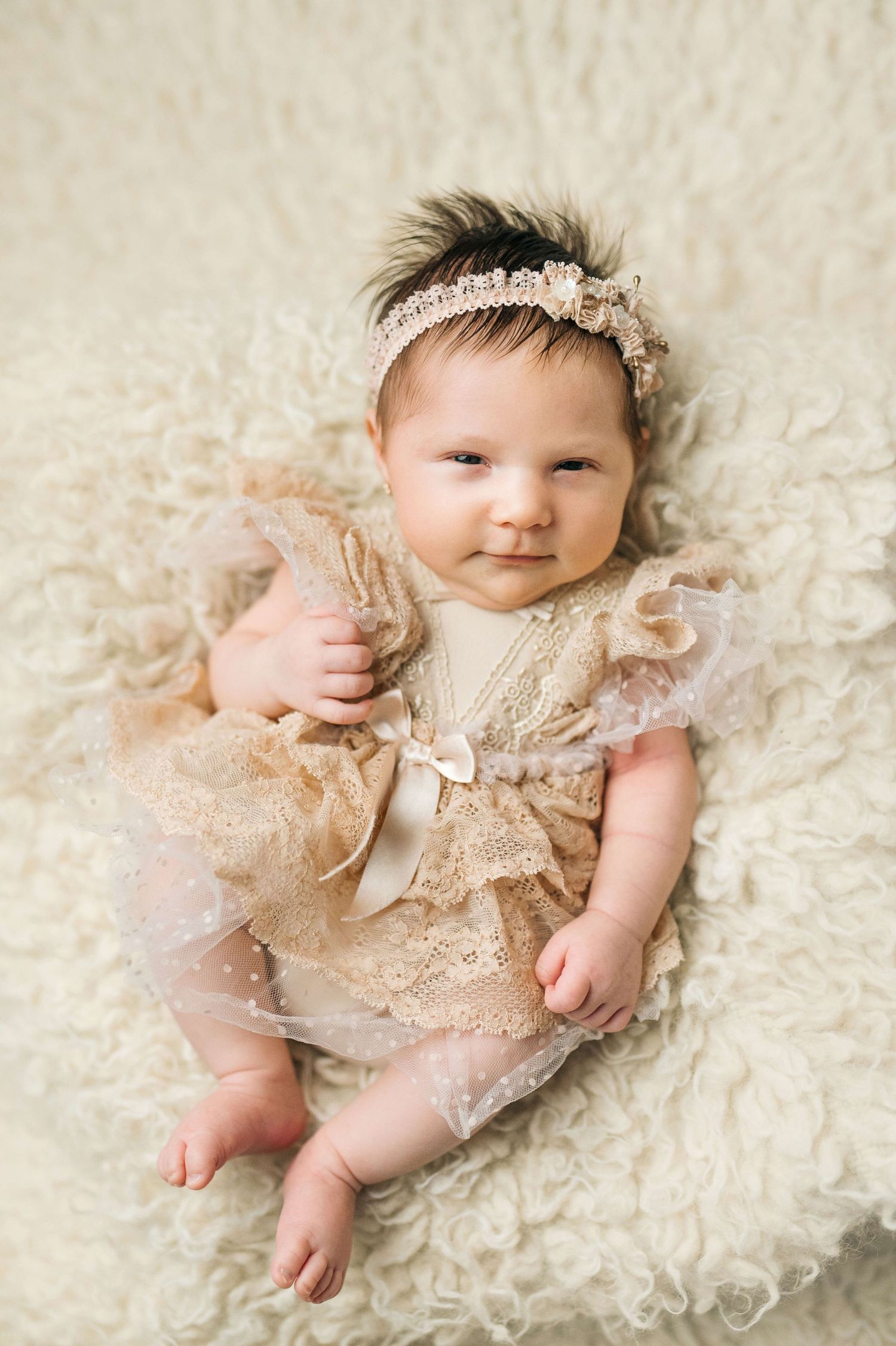 Adorable newborn baby girl in a beige lace dress and headband lying on a fluffy cream blanket.