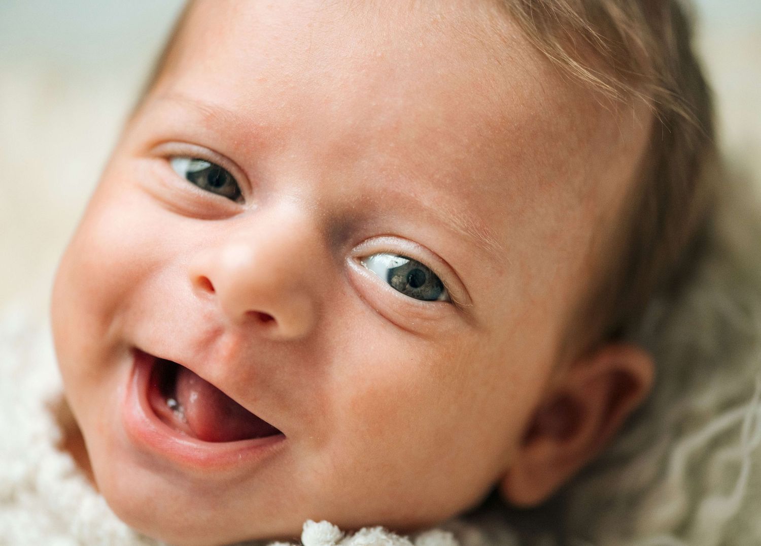 Smiling newborn baby with bright blue eyes lying on a soft white blanket, showing a joyful expression.
