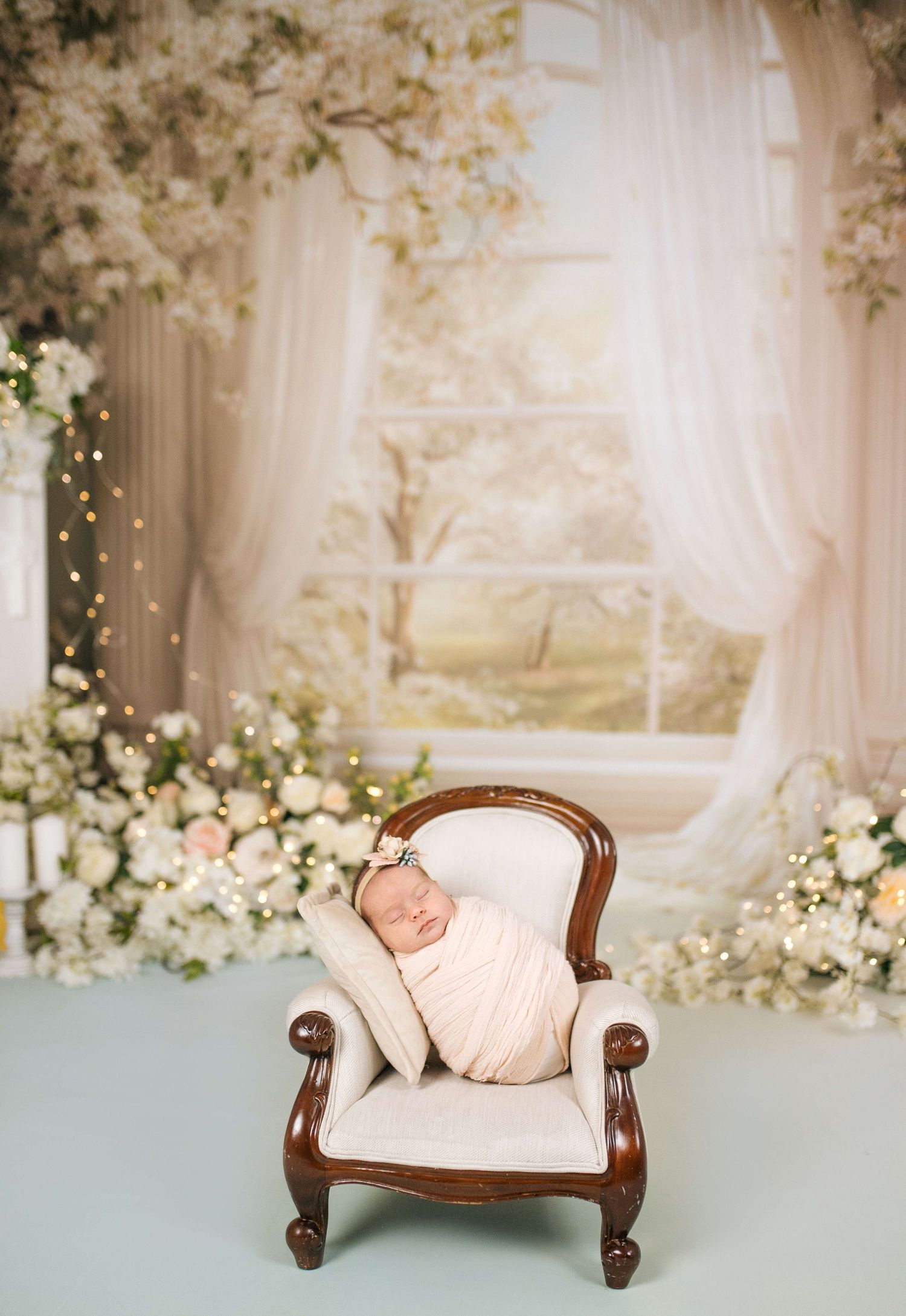 Newborn baby swaddled in cream wrap sleeping peacefully on a miniature armchair surrounded by floral backdrop.