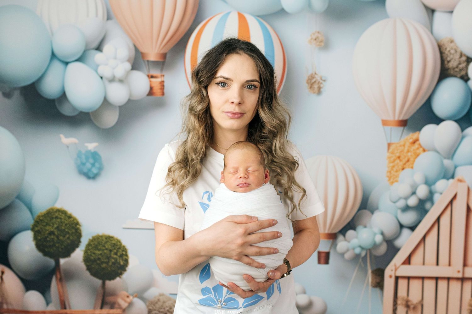 Mother holding newborn baby in front of pastel balloon and hot air balloon decorated backdrop.