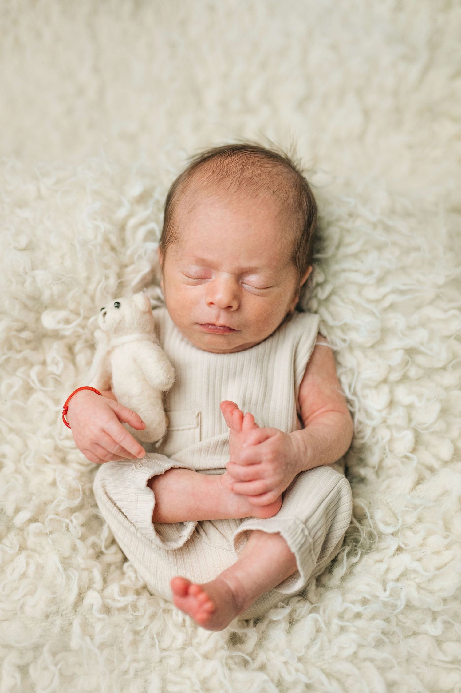 Sleeping newborn baby in cream romper cuddling a small stuffed animal toy on a fluffy white blanket.