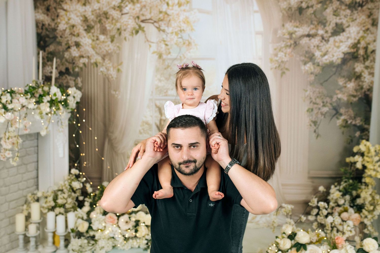 A happy family poses together, father holding toddler on shoulders, mother standing close, surrounded by white floral decor.