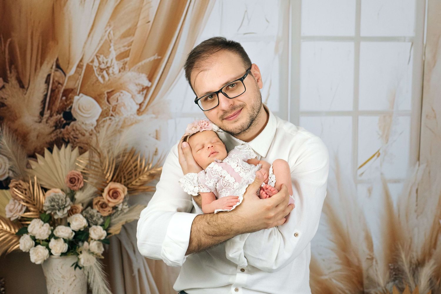 A smiling man in a white shirt wearing glasses gently holds a sleeping newborn baby in a cozy decorated studio.