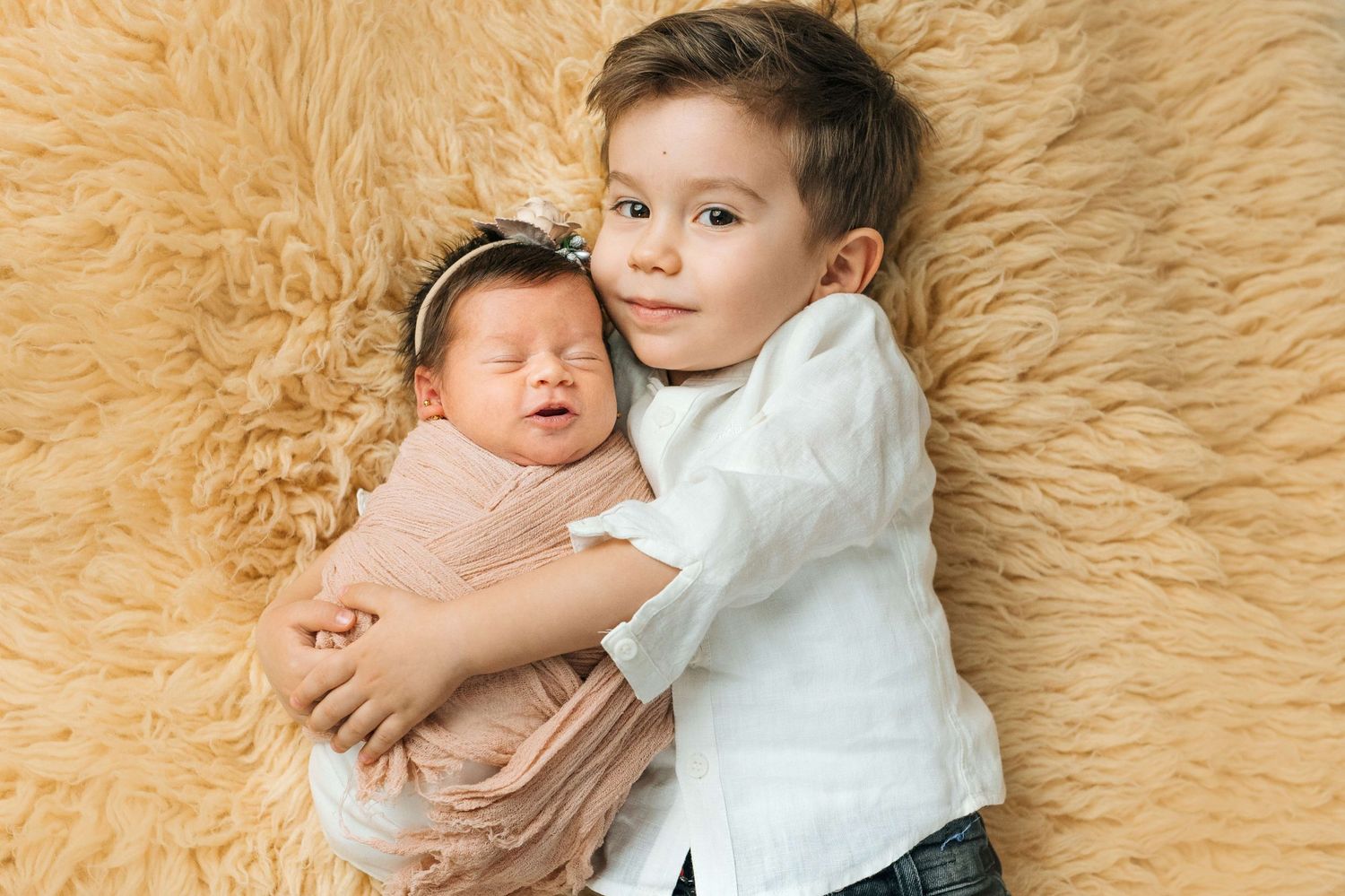 Young boy in white shirt gently holds sleeping newborn baby wrapped in pink on a fluffy beige background.