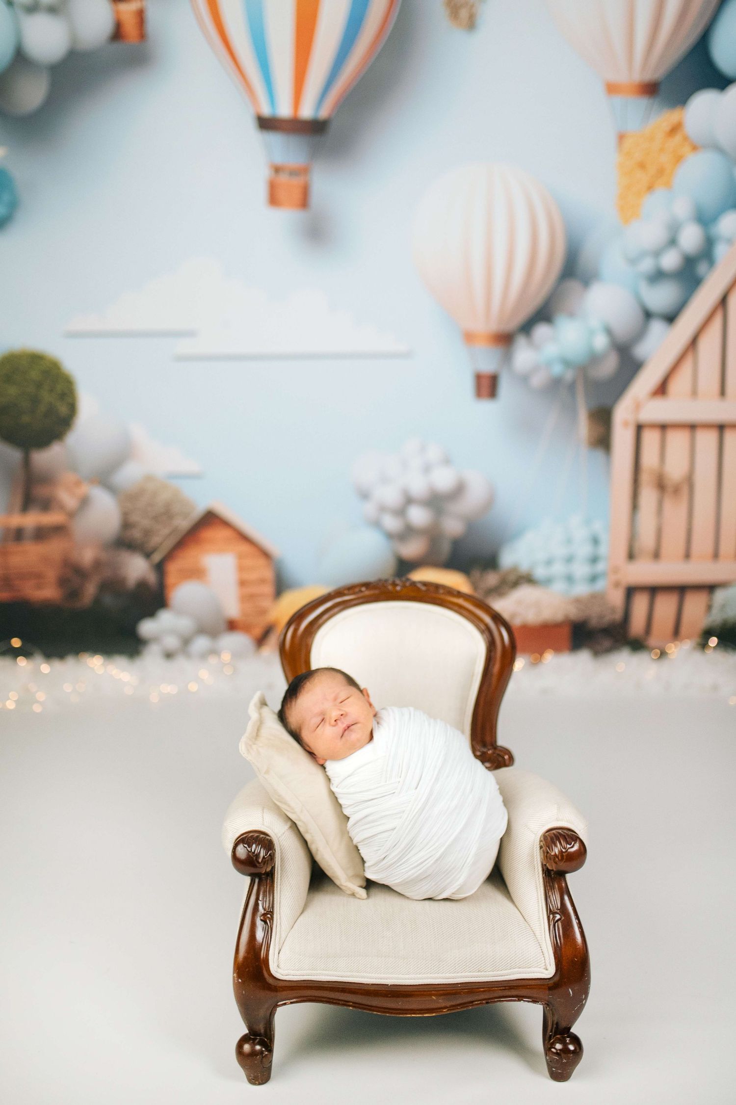 Newborn baby swaddled in white wrap sleeping peacefully in a miniature armchair with hot air balloon nursery backdrop.