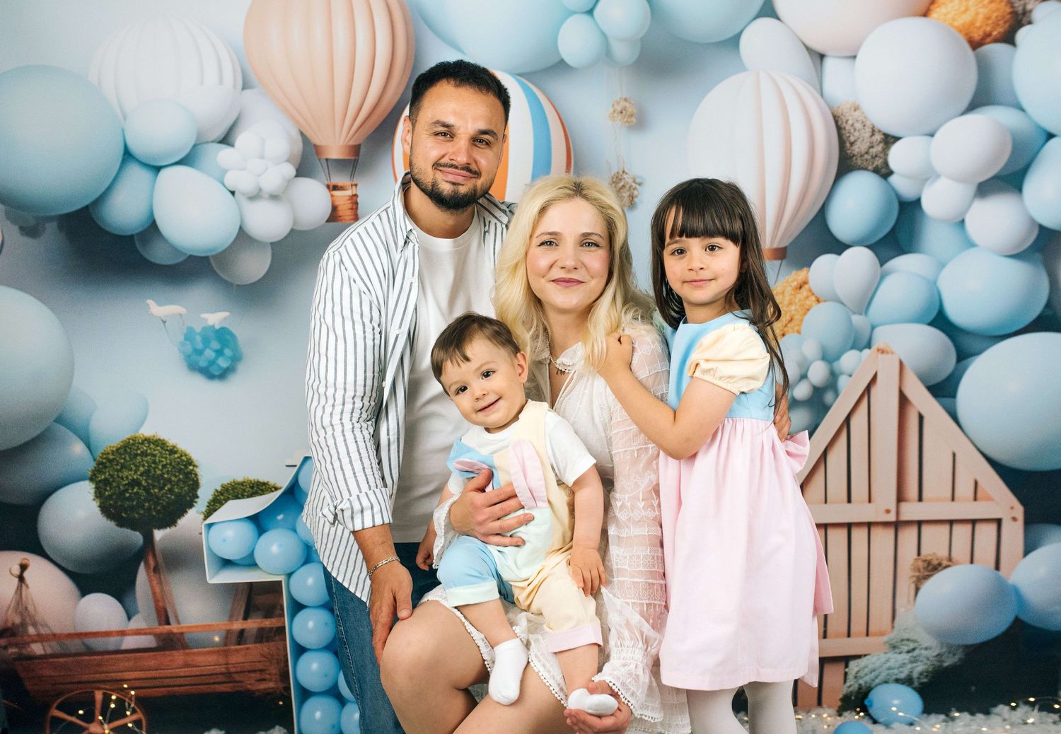 Family of four posing in front of blue balloon backdrop with hot air balloons, parents with toddler and young girl.