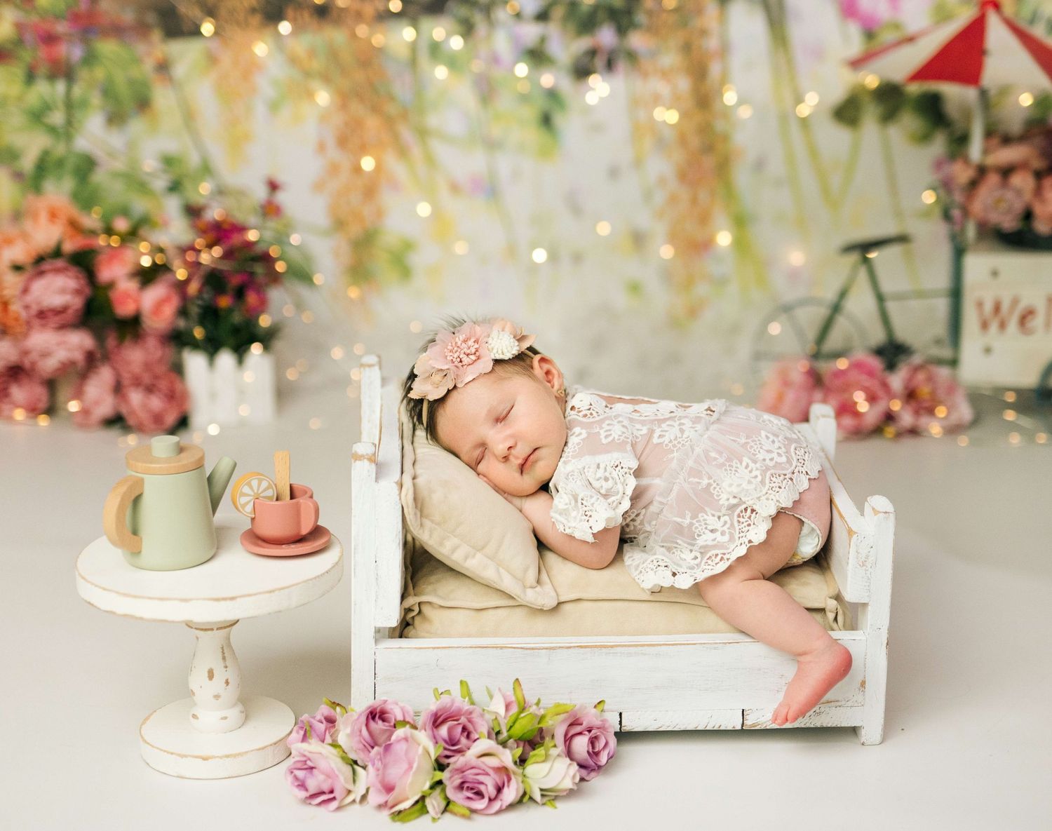Newborn baby sleeping on tiny white bed with floral headband, surrounded by pink roses and bokeh lights.