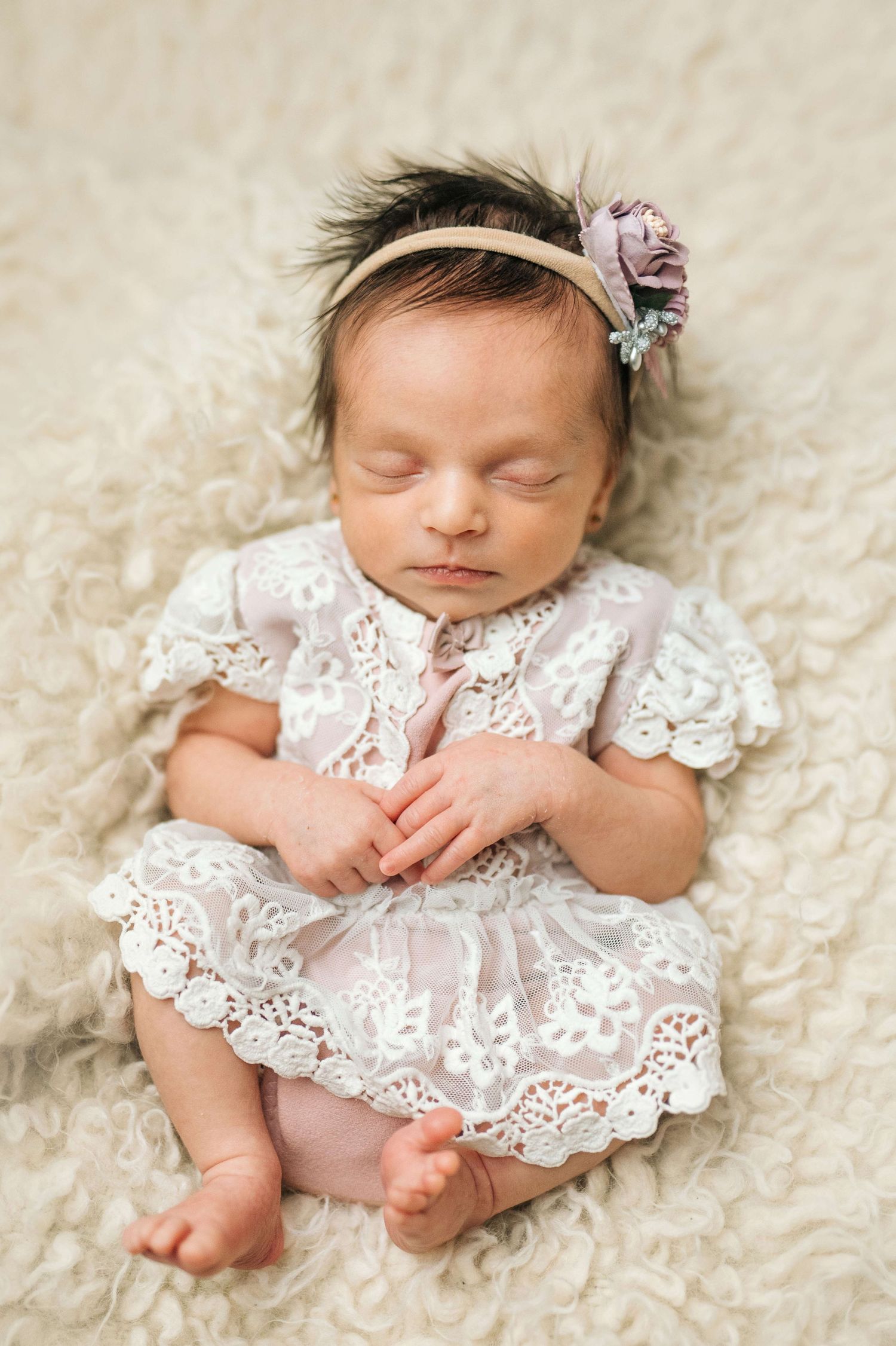 Sleeping newborn baby girl in white lace dress and floral headband on cream fuzzy blanket.