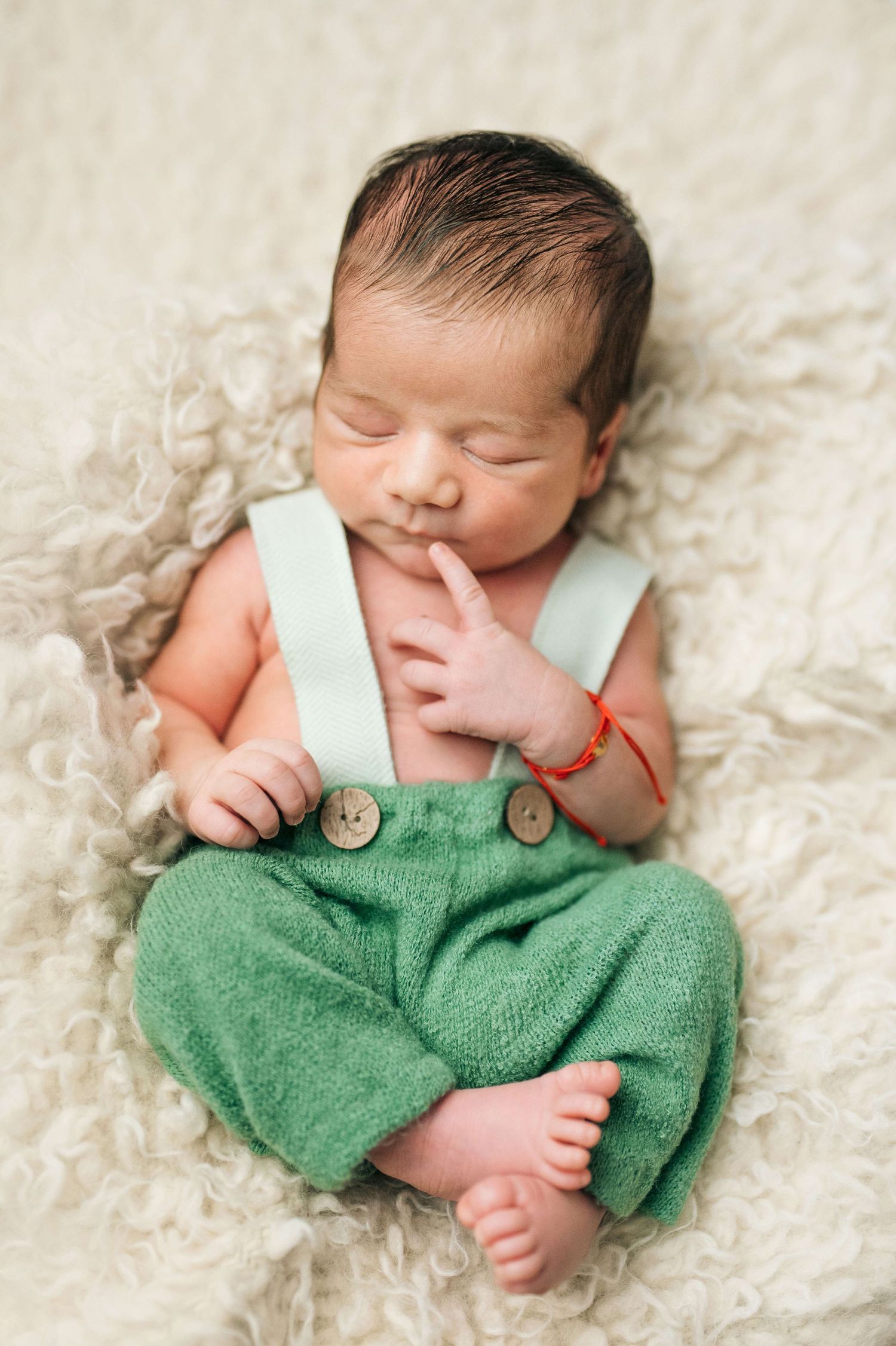 Sleeping newborn baby wearing green suspender pants lying on a fluffy cream blanket with finger near mouth.