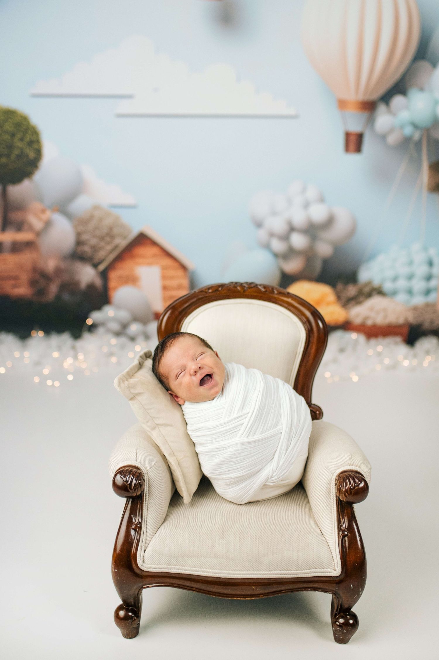 Smiling newborn swaddled in white wrap, posed in miniature armchair with hot air balloon nursery backdrop.