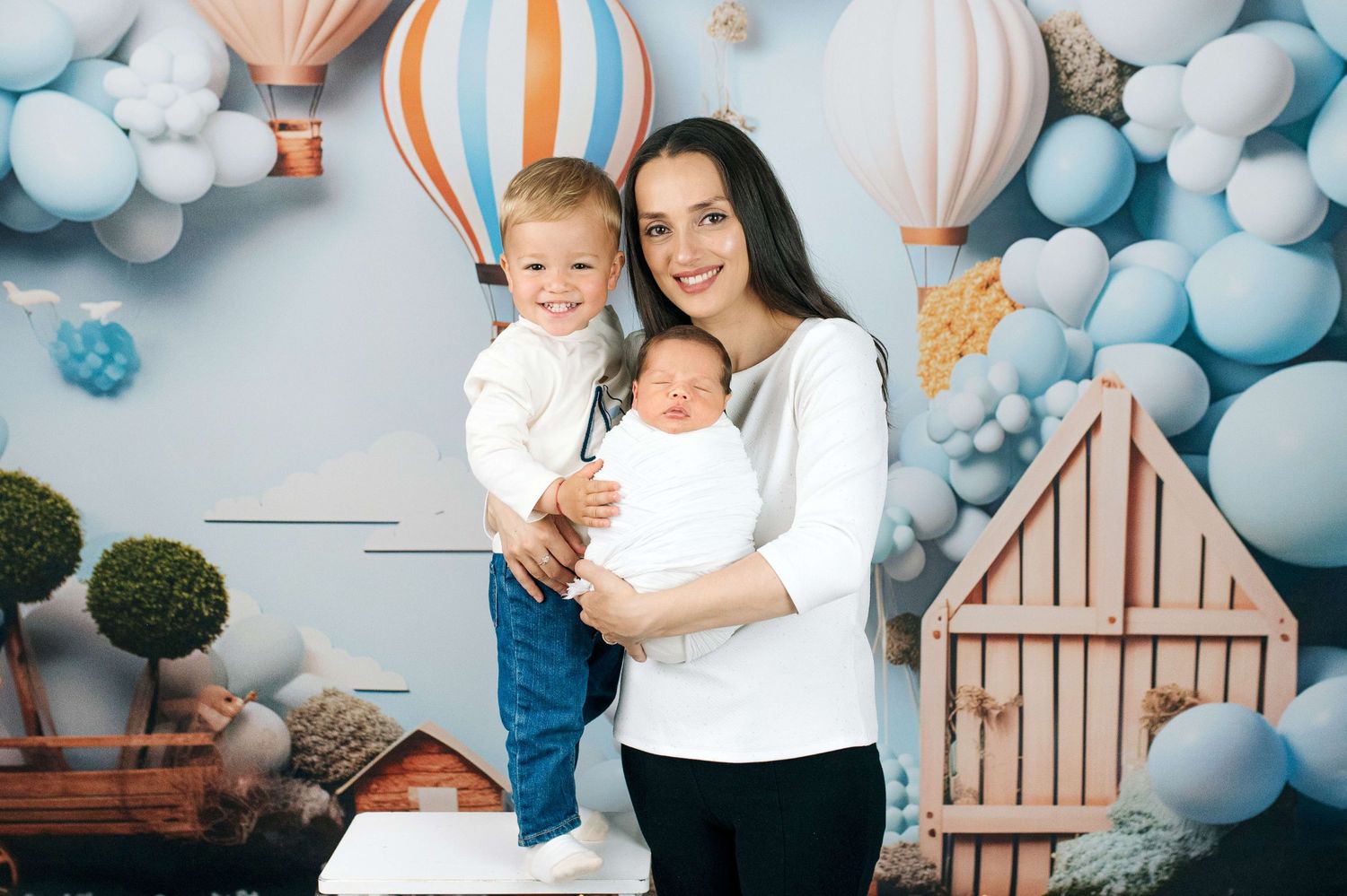 A smiling mother holds a newborn baby while posing with her toddler son in a festive balloon-themed photo studio.
