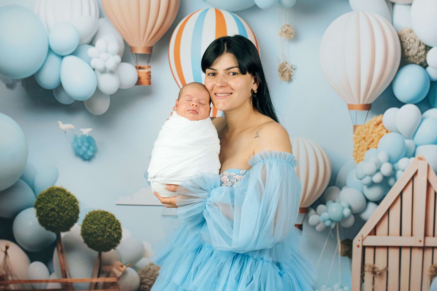 A young mother in a blue tulle dress holds her newborn baby in a dreamy balloon-themed photo shoot.