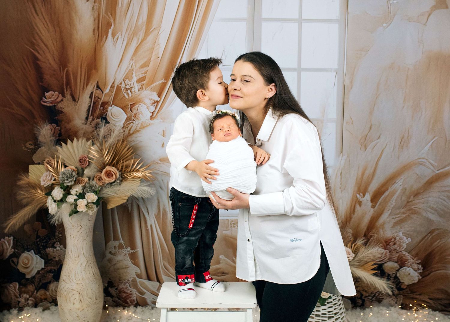 A mother in white holds a newborn while an older child kisses her cheek, surrounded by floral decor.