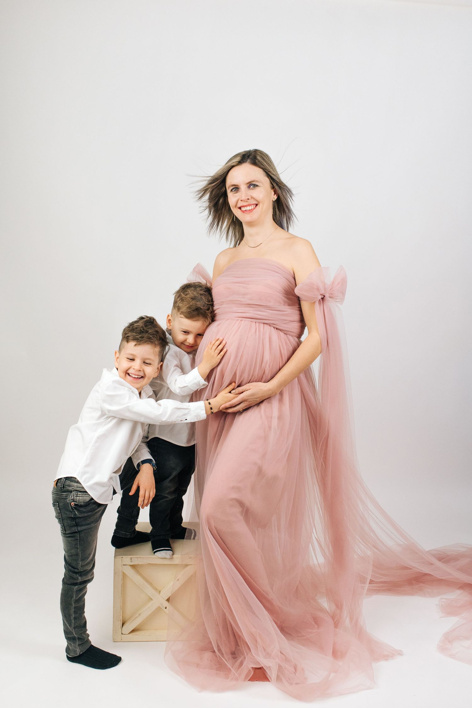 Pregnant woman in pink tulle gown smiling with two young boys hugging her bump in a white studio.