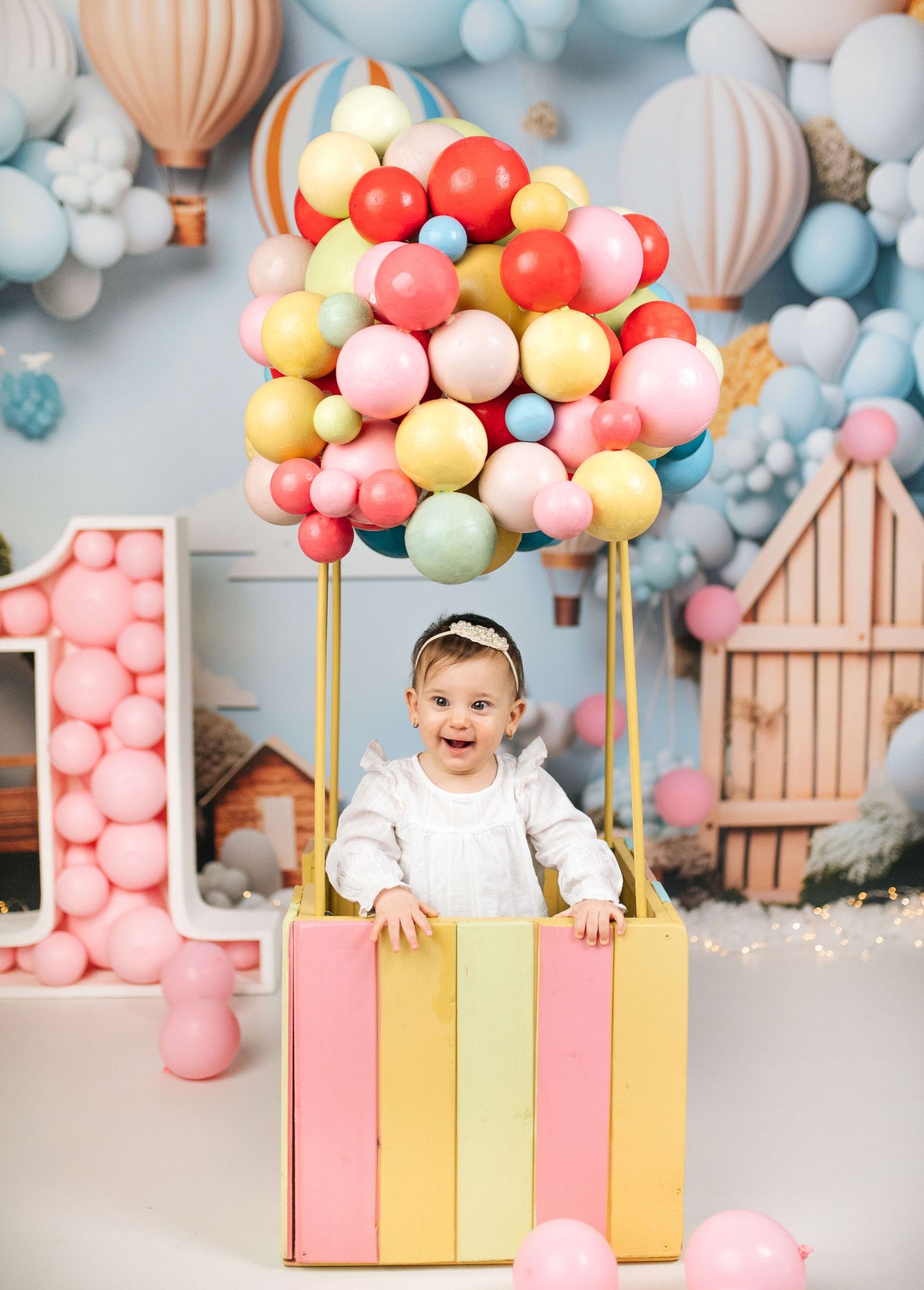 Baby girl sits in colorful hot air balloon prop surrounded by pastel balloons for a first birthday cake smash photo.