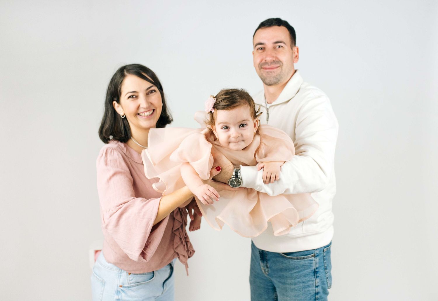 A smiling family of three poses together, with parents holding their toddler daughter in a pink tutu dress.