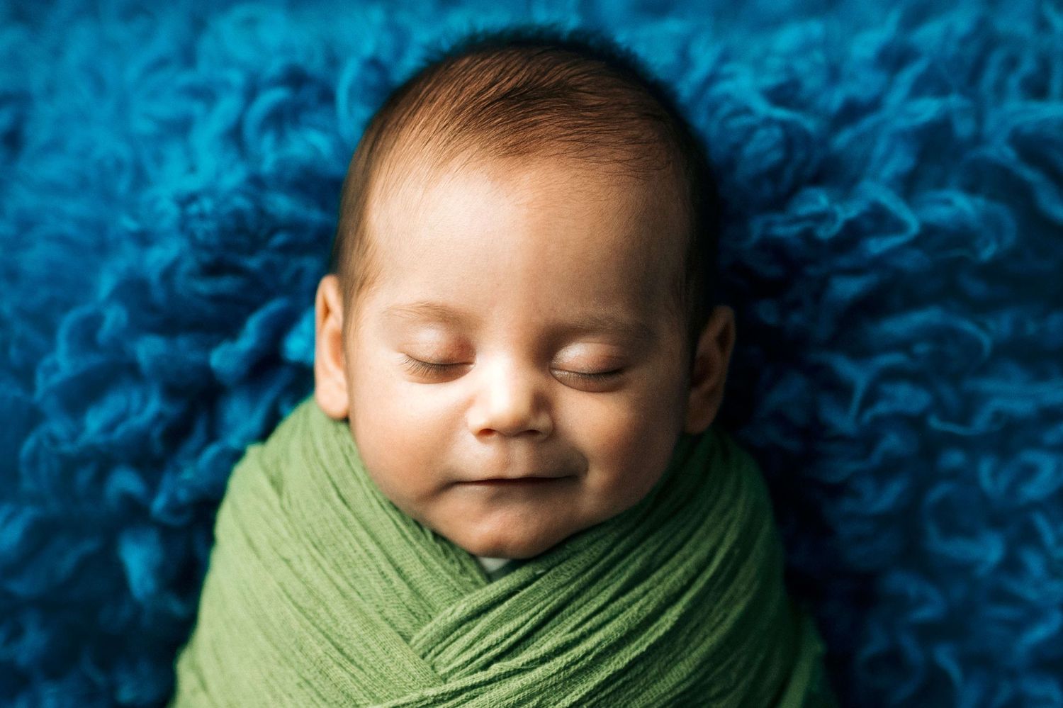 Sleeping newborn baby swaddled in green wrap on blue fluffy blanket, peaceful infant portrait photography.